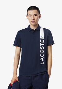 Granatowa koszulka polo z białym pionowym paskiem z napisem "LACOSTE". Krótkie rękawy i klasyczny kołnierzyk. Logo krokodyla Lacoste na piersi.