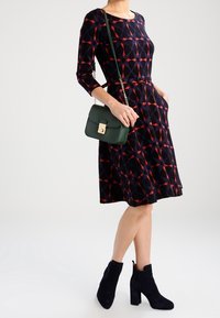 Kvinna klädd i en knälång klänning med geometriskt mönster i marinblått och rött, svarta ankelskor och bärandes en mörkgrön axelremsväska.