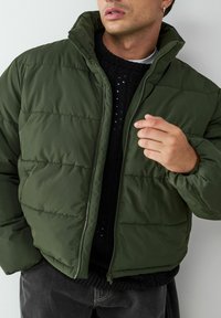 Grüne Puffjacke mit hohem Kragen und Reißverschluss, ausgestattet mit gesteppten Linien und einer glatten Textur, getragen über einem schwarzen Strickpullover.