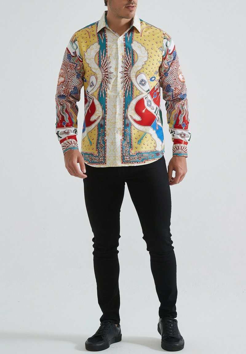 Homme portant un pantalon noir, des baskets noires et une chemise à manches longues à motifs colorés avec des motifs de soleil et abstraits.