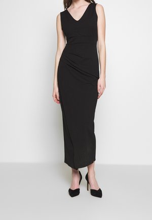 Femme portant une longue robe noire sans manches avec un décolleté en V et des chaussures à talons hauts noires, debout devant un fond clair uni.