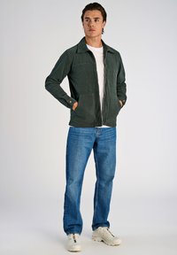 Donkergroen zip-up jack met een kraag en twee voorzakken, gecombineerd met lichtblauwe jeans en witte sneakers. De stof heeft een gestructureerde afwerking.