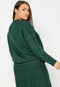 Suéter verde de punto con un patrón texturizado de cable en las mangas y el dobladillo, con un corte relajado y puños acanalados.