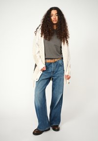 Weite blaue Jeans kombiniert mit einem grauen T-Shirt und einer cremefarbenen Strickjacke mit Zickzackmuster. Dunkle Schuhe runden den Look ab.