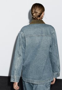 Persoon met lichtbruin haar in een bun, gekleed in een lichtblauwe spijkerjack met een bruine kraag, staand tegen een witte muur.