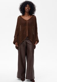 Brązowy sweter na szydełku o luźnym kroju, szerokich rękawach i głębokim dekolcie w kształcie litery V, zestawiony z brązowymi skórzanymi spodniami o wysokim stanie i spiczastymi obcasami.