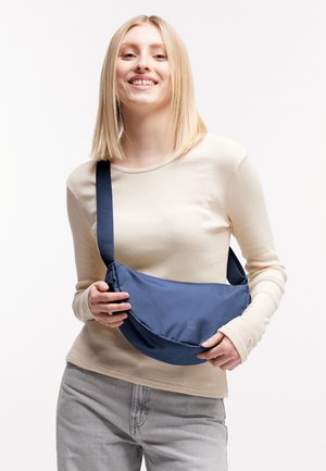 Mujer sonriente con blusa beige de manga larga y pantalones grises, sosteniendo y llevando un bolso bandolera azul con correa ajustable.