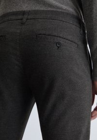 Donkergrijze tekstuurbroek met een subtiel gestreept patroon, voorzien van een achterzak met een zwarte knop en een geribde tailleband.