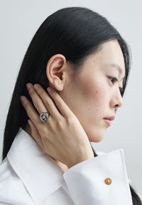 Ezüst gyűrű, nagy, kiemelkedő középső kővel, kisebb kövekkel körülvéve, egy semleges bőrtónusú kézen viselve, világos háttér előtt.