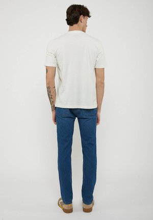 Man met getatoeëerde linkerarm draagt wit T-shirt met korte mouwen, blauwe spijkerbroek en beige schoenen, staand met gezicht naar een witte achtergrond.