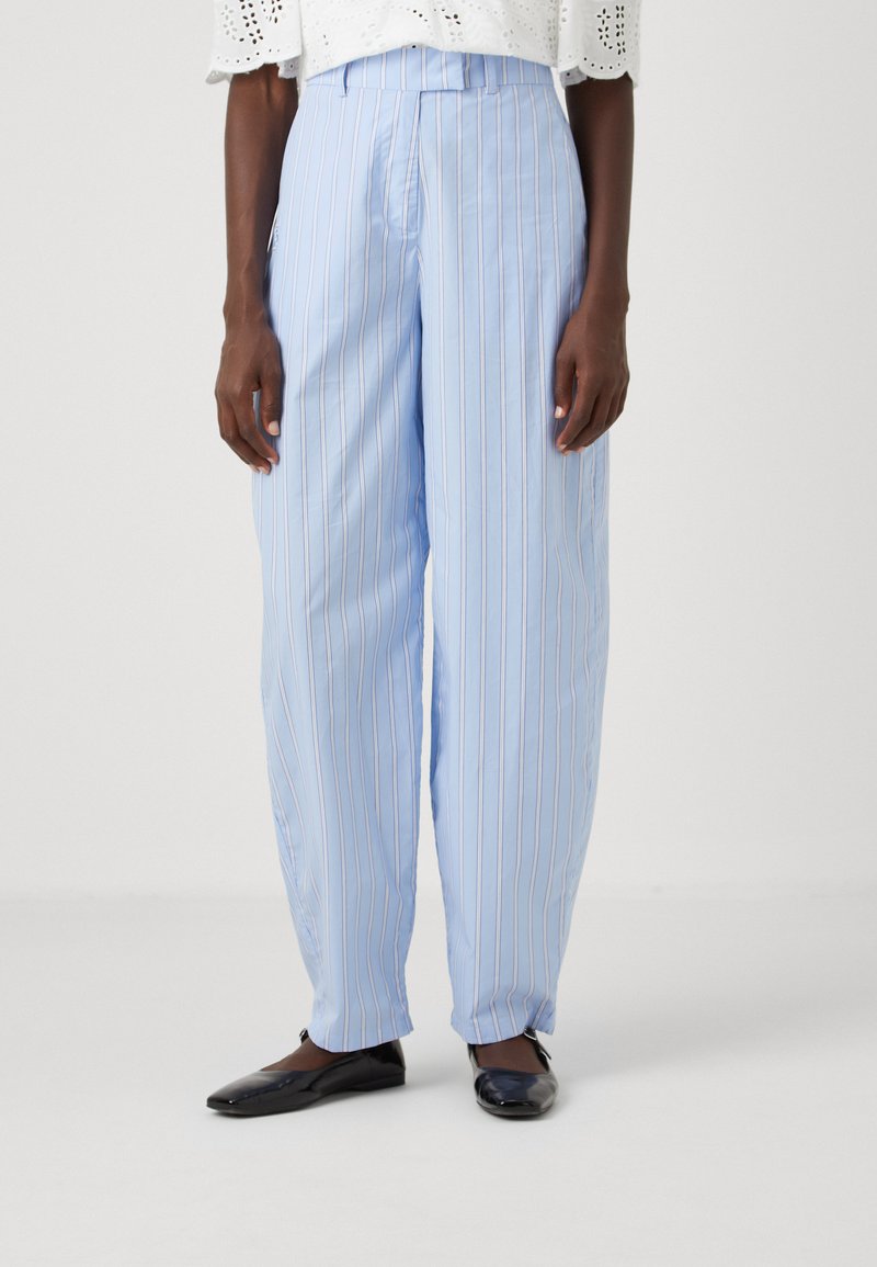 Lichtblauwe wijde broek met verticale witte strepen, op maat gemaakte tailleband en voorplooien. G gedragen met zwarte platte schoenen.