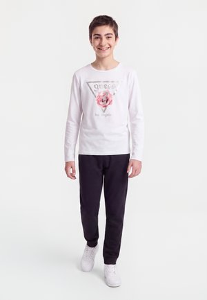 Garçon souriant marchant en avant portant une chemise blanche à manches longues avec un motif de rose rose et un pantalon noir avec des baskets blanches.