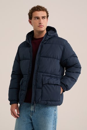 Homme portant une veste matelassée à capuche bleu marine sur un pull sombre et un jean bleu, regardant sur le côté avec une main dans sa poche.