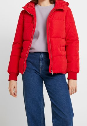 Personne portant une doudoune rouge par-dessus un pull gris clair et un jean bleu, debout devant un fond clair uni.