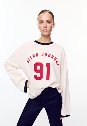 Donna con capelli tirati indietro indossa una felpa crema larga con la scritta "FLEUR JOURNAL 91" in rosso e polsini rifiniti in nero, abbinata a pantaloni scuri.