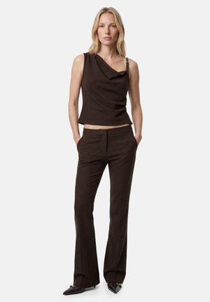 Femme aux cheveux blonds portant un top asymétrique sans manches marron foncé et un pantalon tailleur assorti avec des chaussures à bouts pointus, debout les mains dans les poches.