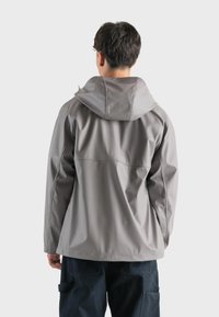 Giacca impermeabile grigia con cappuccio, caratterizzata da una texture liscia e cuciture visibili, abbinata a pantaloni cargo scuri. Vista da dietro.