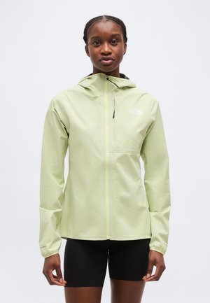 Woman wearing light green hooded jacket zipped up, standing with arms relaxed, paired with black cycling shorts, against plain background.