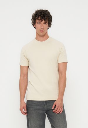 Giovane uomo con capelli ricci che indossa una semplice maglietta beige a maniche corte e jeans grigio scuro, in piedi davanti a uno sfondo bianco.
