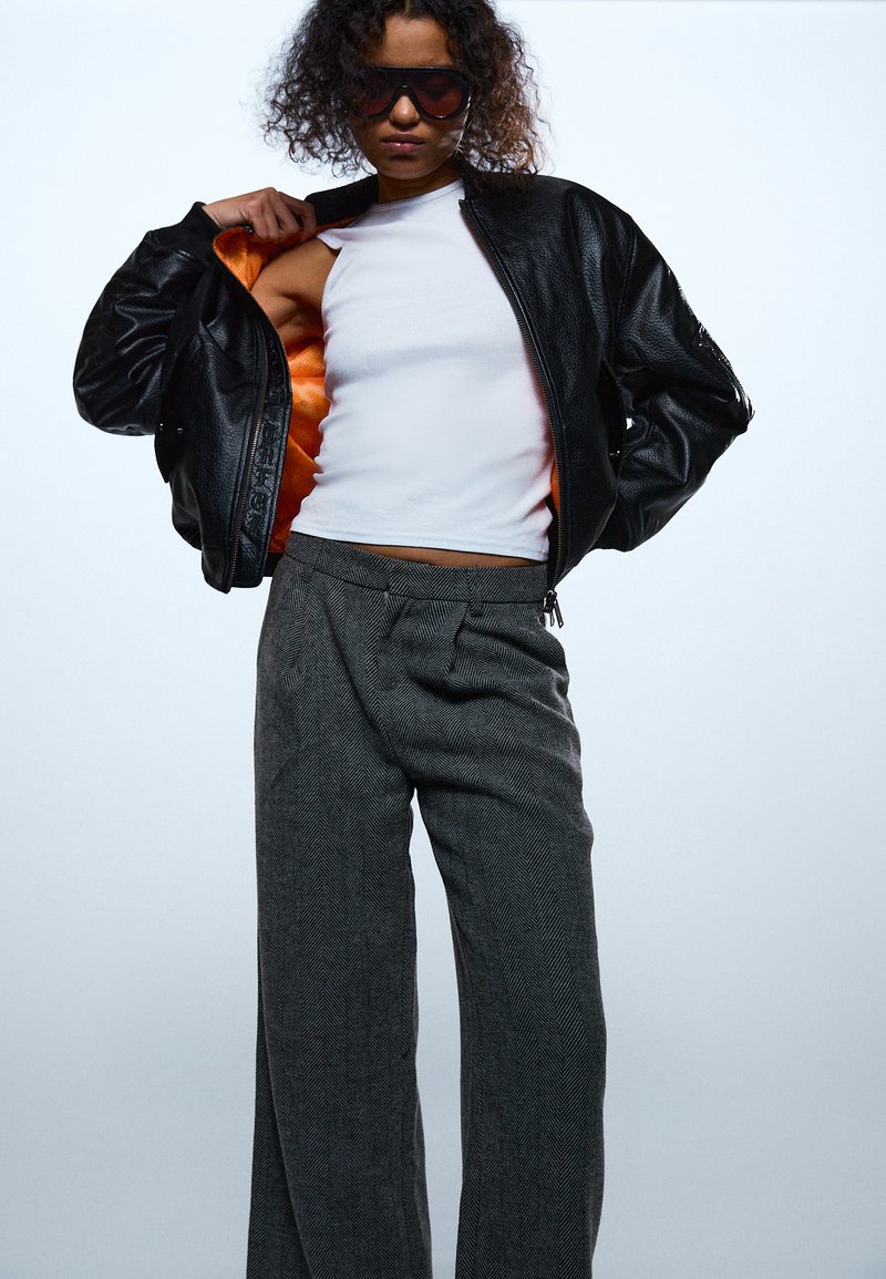 Oseba z velikimi črnimi sončnimi očali, odprto črno usnjeno jakno z oranžno podlogo, belo zgornjo brez rokavov in sivimi hlačami širokih nogavic.