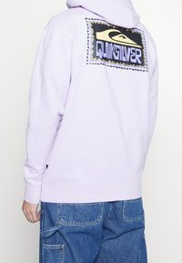 Lila hoodie med en grafisk logotyp på baksidan, som har starka färger och en zigzag-kant. Tyget är mjukt, med en avslappnad passform och ribbade ärmmuddar.