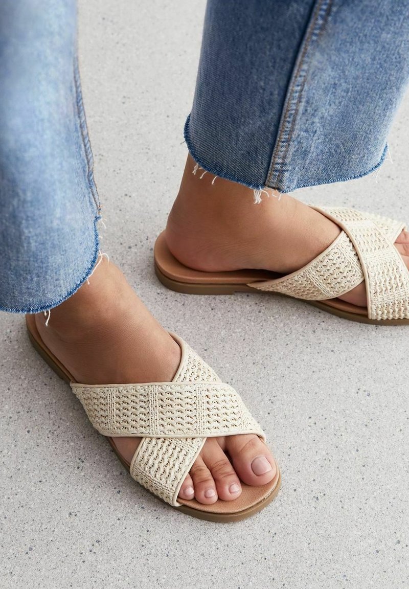 Pieds portant des sandales beige tissées à brides croisées et un jean bleu à ourlet effiloché sur un sol texturé gris clair.