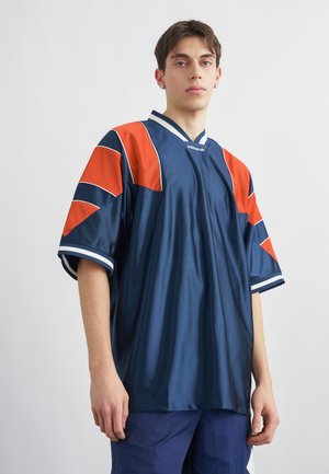 Νεαρός άνδρας που φοράει άνετη αθλητική φανέλα Adidas σε μπλε ναυτικό και πορτοκαλί χρώμα με κοντά μανίκια, στέκεται μπροστά σε απλό φωτεινό φόντο.