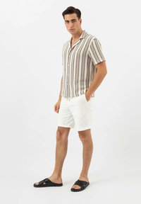 Chemise à manches courtes, boutonnée, avec des rayures verticales beiges et noires ; assortie à un short blanc et des sandales noires à glissière.