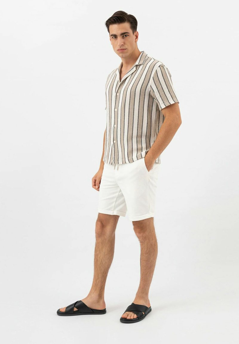 Chemise à manches courtes, boutonnée, avec des rayures verticales beiges et noires ; assortie à un short blanc et des sandales noires à glissière.