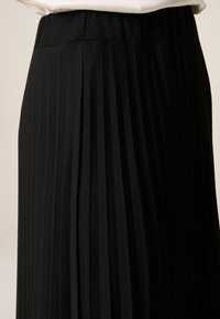 Musta laskostettu hame, jossa on kohokuvioitu kangas, pystysuuntaiset raidat ja joustava vyötärönauha mukavuutta ja helppoa pukeutumista varten.