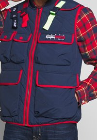 Blå quiltad väst med röda accenter, flera framfickor och en logotypapplikation. Har gröna och svarta remmar på axlarna.