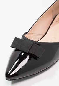 Chaussure plate noire brillante à bout pointu avec un nœud en tissu noir texturé sur la tige et une doublure intérieure beige.