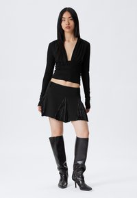 Woman with long black hair wearing a deep V-neck black long-sleeve top, black mini skirt with pleats, and knee-high black leather boots.