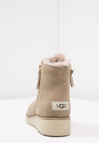 Botín de ante marrón con forro de felpa, cremallera dorada y etiqueta de logo. Presenta una suela de goma texturizada para agarre y estabilidad.