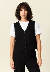 Svart väst med V-ringning, med sex knappar, två framfickor och en figurnära passform. Bärs över en vit t-shirt.