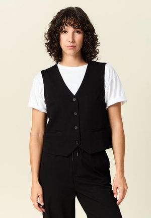 Chaleco negro con un diseño de escote en V, que cuenta con seis botones, dos bolsillos delanteros y una forma ajustada. Lleva puesto sobre una camiseta blanca.