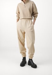 Beige cropped sweatshirt met kwart ritssluiting en logo, gecombineerd met crèmekleurige broek met een geometrisch patroon en elastische tailleband, gedragen met zwarte laarzen.