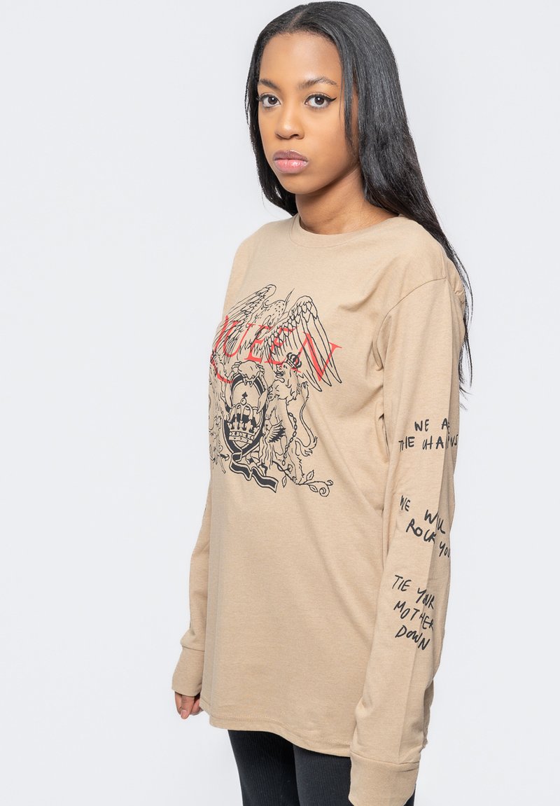 Beige long-sleeve t-shirt with graphic print featuring wings and a crown. Text details on sleeves; soft fabric and relaxed fit.