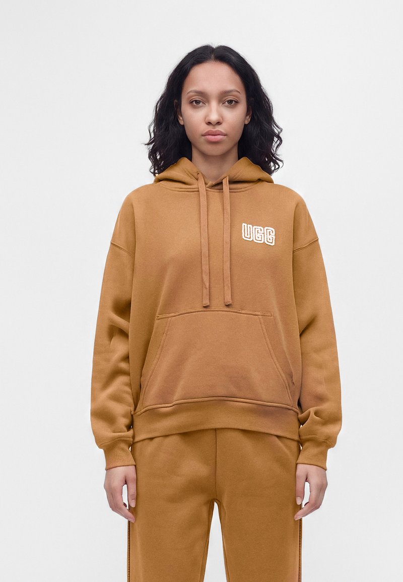 Jeune femme aux cheveux bouclés et foncés portant un sweat à capuche marron UGG et un pantalon assorti, debout droite devant un fond clair uni.