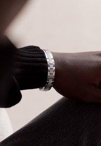 Bracelet en argent avec des maillons entrelacés, présentant un bandeau texturé et des accents lisses, porté sur un poignet à la peau foncée.