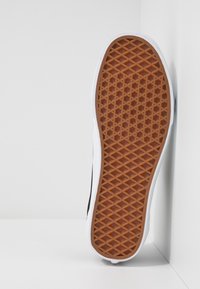 Hvit sneakersåle med en brun gummibunn som har et diamantmønster, svarte sideaksenter og glatte hvite gummikanter.