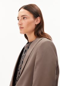 Jonge vrouw met bruin haar in een taupe blazer en een sweater met luipaardprint, die nadenkend naar links kijkt tegen een witte achtergrond.