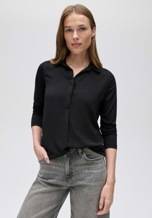 Donna con capelli castano chiaro che indossa una camicetta nera a maniche lunghe e jeans grigi, in piedi con una mano in tasca su uno sfondo uniforme.