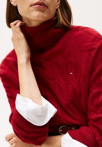 Jersey de cuello alto de punto rojo con patrón de trenzas, combinado con una camisa blanca de cuello que asoma por los puños. Detalle mínimo del logo de la marca.