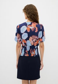 Bluse mit Blumenmuster in Marineblau und Pastelltönen, mit einem enganliegenden, gerafften Bund und kurzen Puffärmeln. Kombiniert mit einem dunklen Rock.
