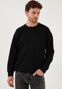 Črna majica s kapuco z okroglim izrezom in raglan rokavi, izdelana iz gladkega materiala. Ima majhen, subtilen logo spredaj.