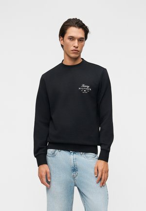 Felnőtt férfi fekete Tommy Hilfiger pulóverben és világoskék farmerral, semleges arckifejezéssel előre nézve.
