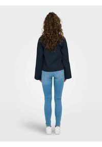 Chaqueta navy de manga larga, diseño ajustado, vista desde atrás. Combinada con jeans skinny azul claro y zapatillas blancas.