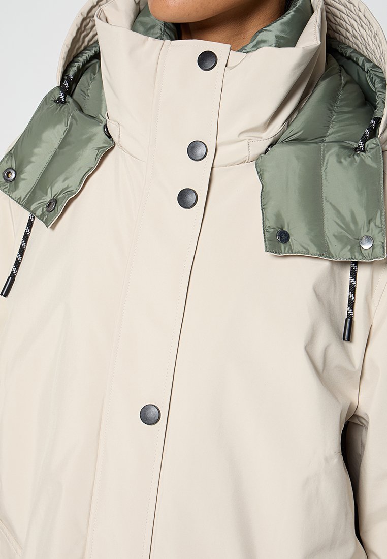 Veste beige à col montant avec boutons pressions noirs et capuche matelassée olive amovible avec cordons noir et blanc.