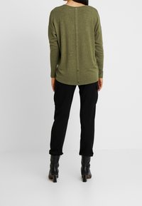 Maglia verde oliva a maniche lunghe con scollo arrotondato, tessuto strutturato e dettagli con bottoni lungo la schiena. Abbinata a pantaloni cargo neri e stivali con tacco.